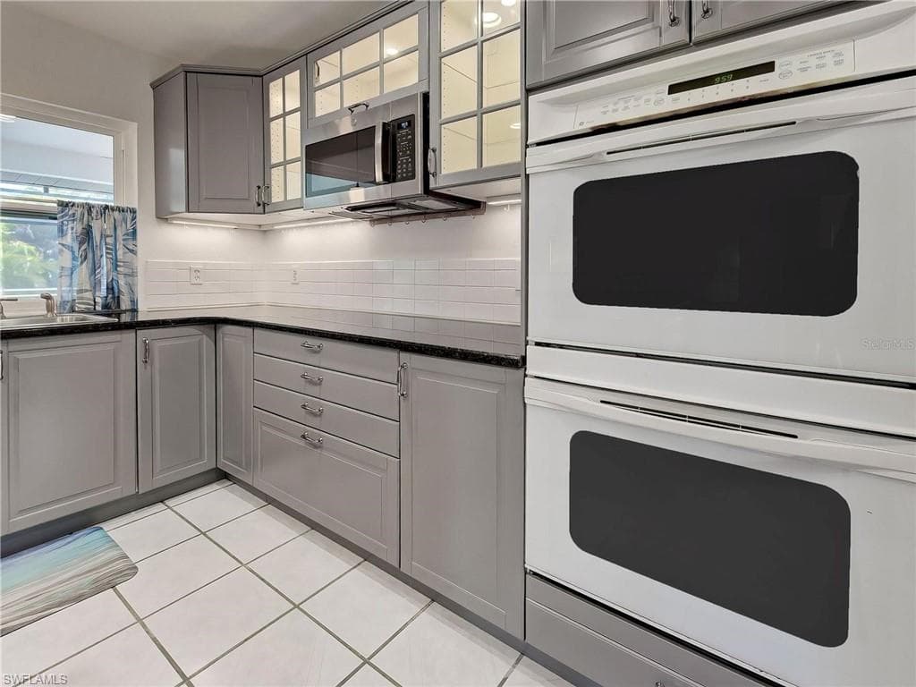 Kitchen with gray cabinetry, white double oven, dark stone countertops, stainless steel microwave, and light tile patterned floors