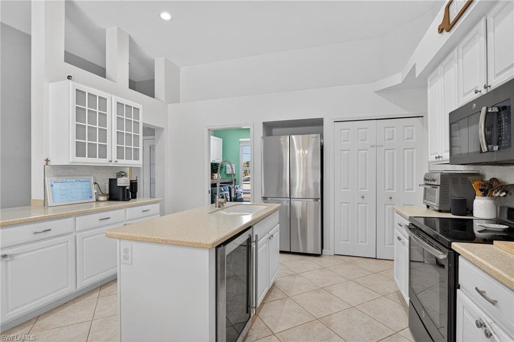 Kitchen featuring stainless steel appliances, white cabinetry, a center island with sink, beverage cooler, and glass insert cabinets
