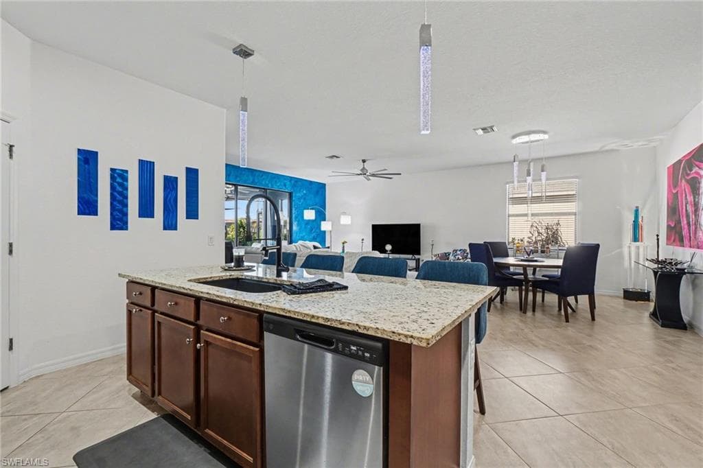 Kitchen featuring stainless steel dishwasher, light stone countertops, pendant lighting, and a center island with sink