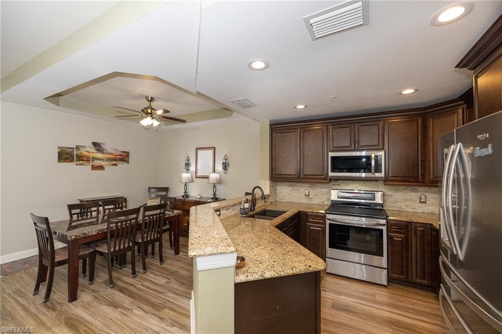 Kitchen featuring dark wood finish cabinetry, stainless steel appliances, a raised ceiling, a ceiling fan, and light stone countertops