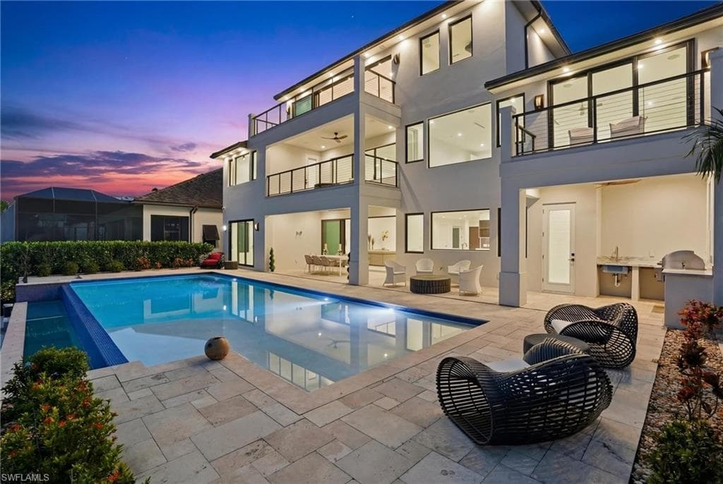 Back of property at dusk featuring stucco siding, a patio area, a balcony, and an infinity pool