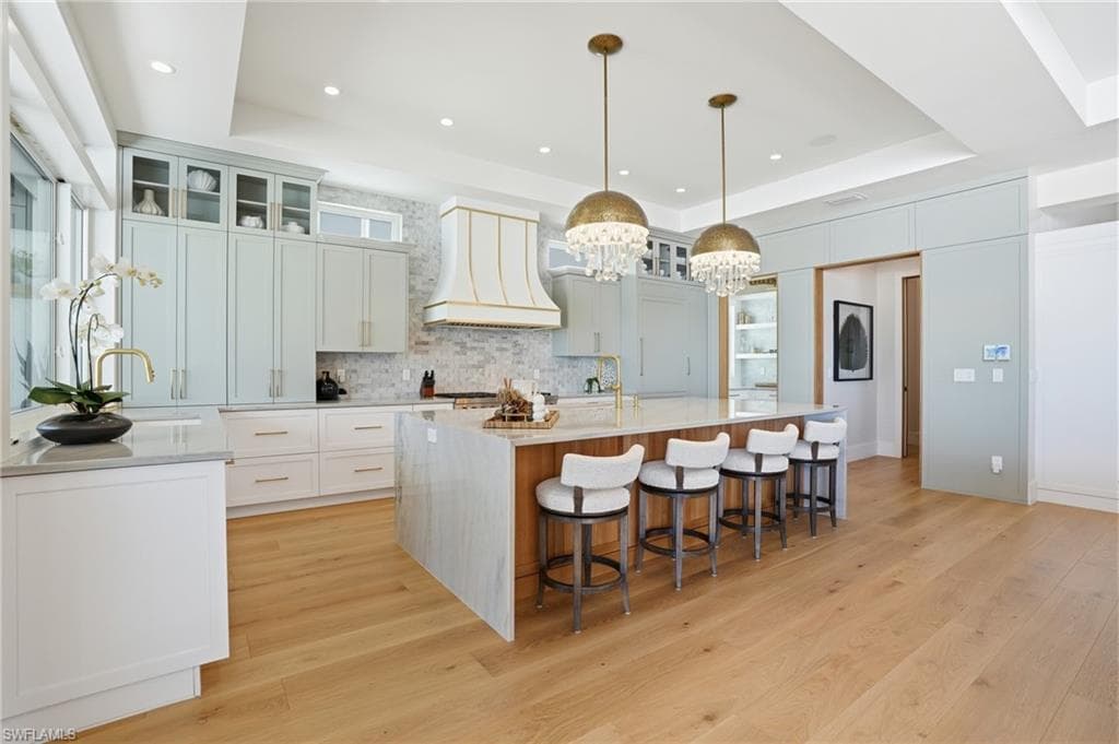Kitchen with glass fronted cabinets, light wood-type flooring, light stone countertops, a large island with sink, and a tray ceiling - Virtually Edited Image