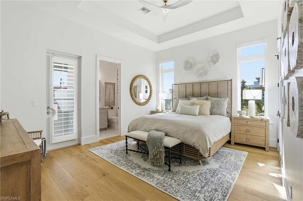 Bedroom with light wood finished floors, ensuite bathroom, a ceiling fan, and a tray ceiling - Virtually Edited Image