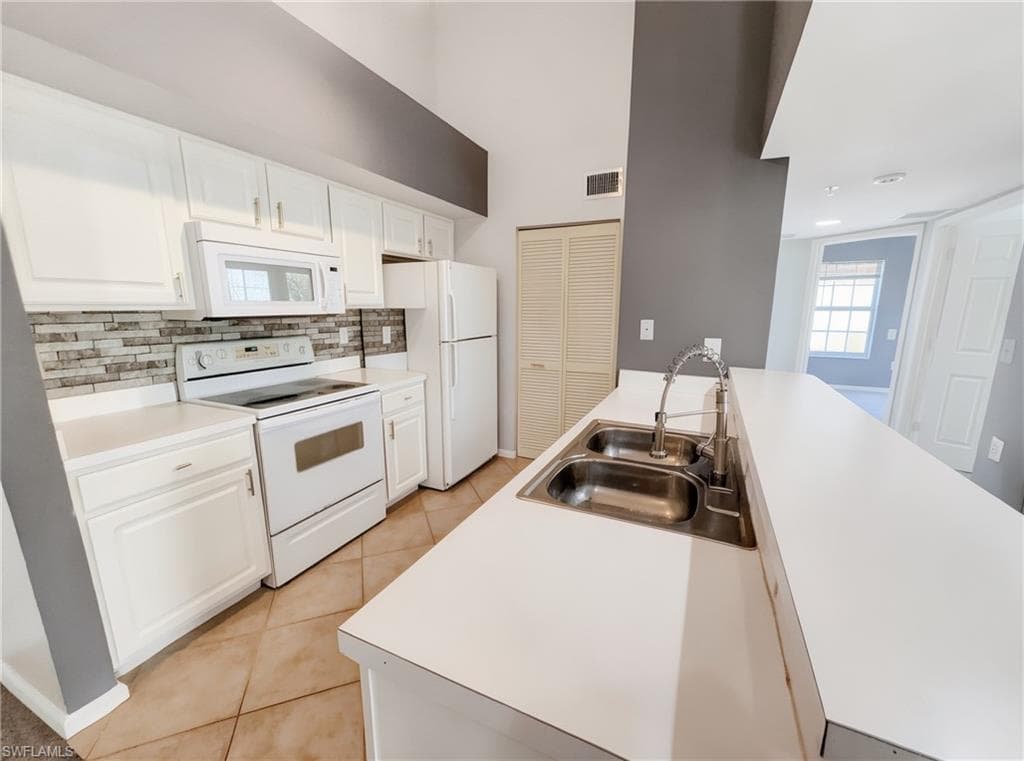 Kitchen featuring white cabinetry, white appliances, a stone-look tile backsplash, a double basin stainless steel sink, and tile flooring