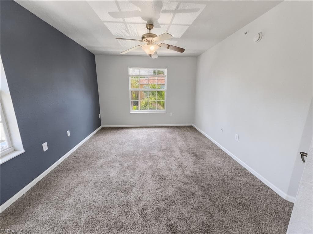 Spacious room featuring plush gray carpeting, a window offering natural light, a ceiling fan with integrated lighting, and a distinct accent wall