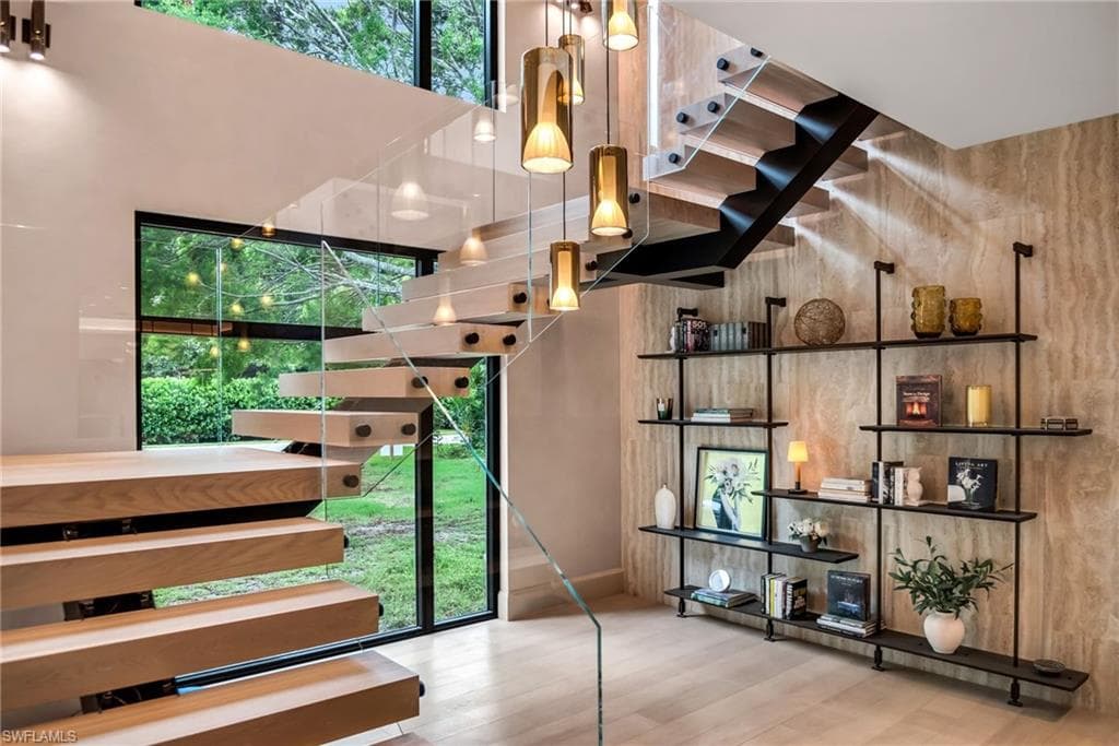 Open tread lit staircase lined on both double-height walls with unfilled travertine and featuring an imported Italian leaning bookshelf below