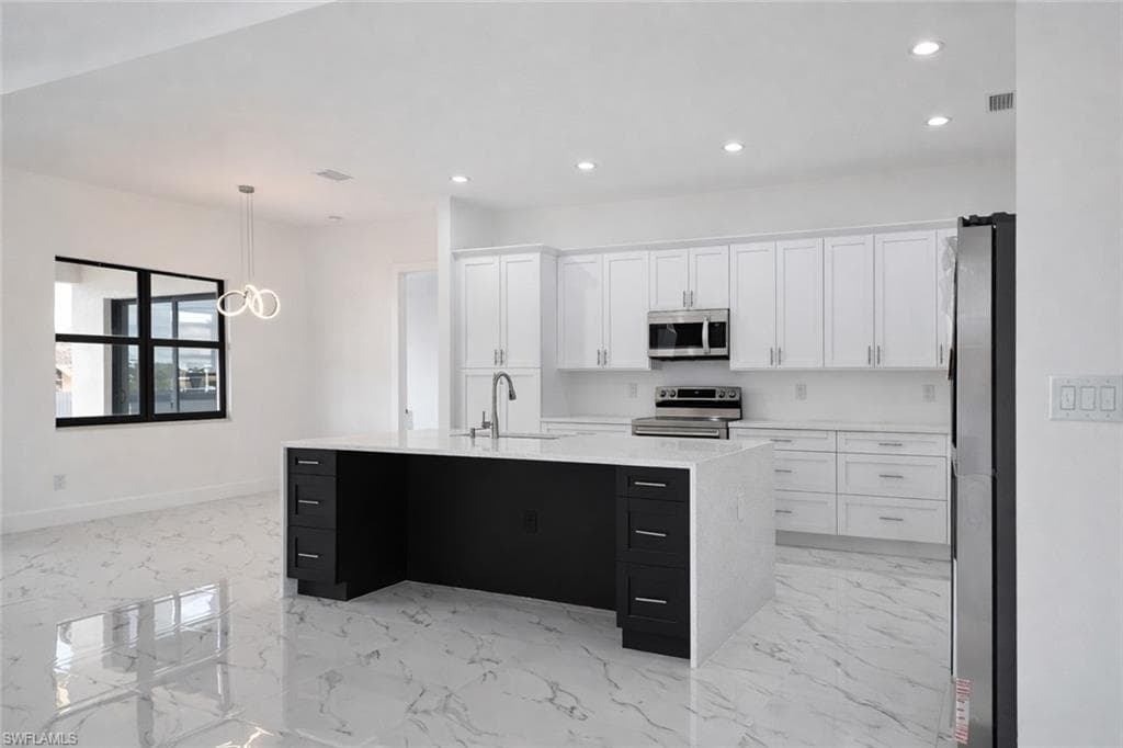 Dual tone kitchen with stainless steel appliances, light stone counters, a kitchen island with sink, hanging light fixtures, and light marble finish flooring