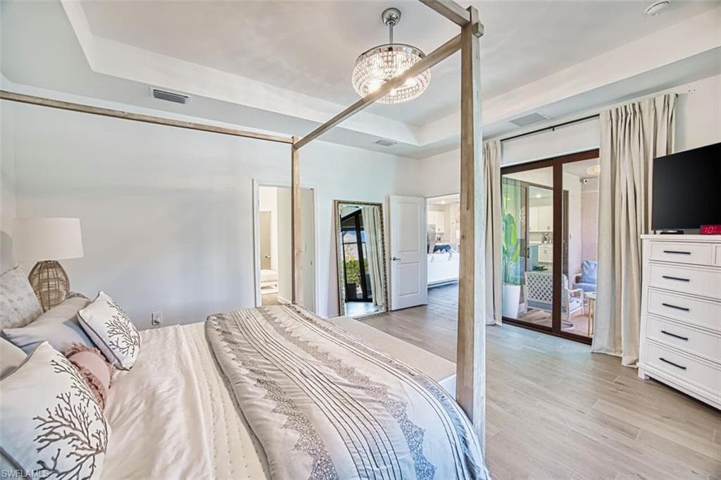 Bedroom with access to outside, light wood-type flooring, a raised ceiling, ensuite bathroom, and suspended lighting