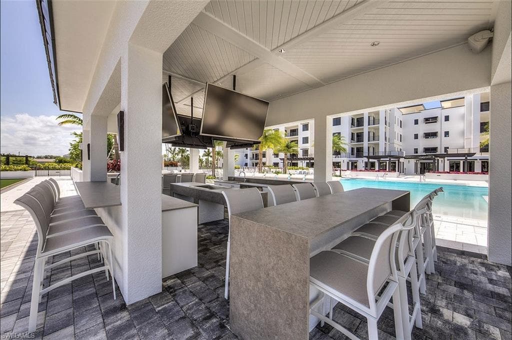 Covered outdoor entertaining area featuring multiple built-in bars with stone-finish countertops, overhead ceiling fans, built-in televisions, and textured paver flooring