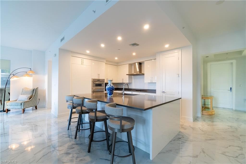 Kitchen featuring white cabinets, a breakfast bar area, a center island with sink, light marble finish floors, and recessed lighting