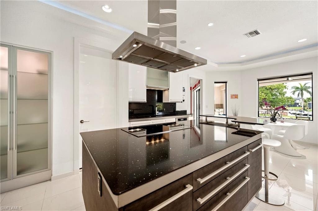Two tone kitchen featuring range hood, modern cabinets, dark stone countertops, recessed lighting, and a breakfast bar area