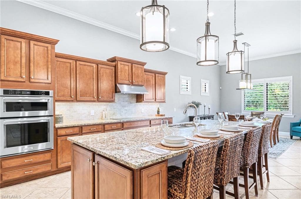Kitchen featuring wood finish cabinets, a kitchen breakfast bar, a kitchen island with sink, light stone counters, and ornamental molding