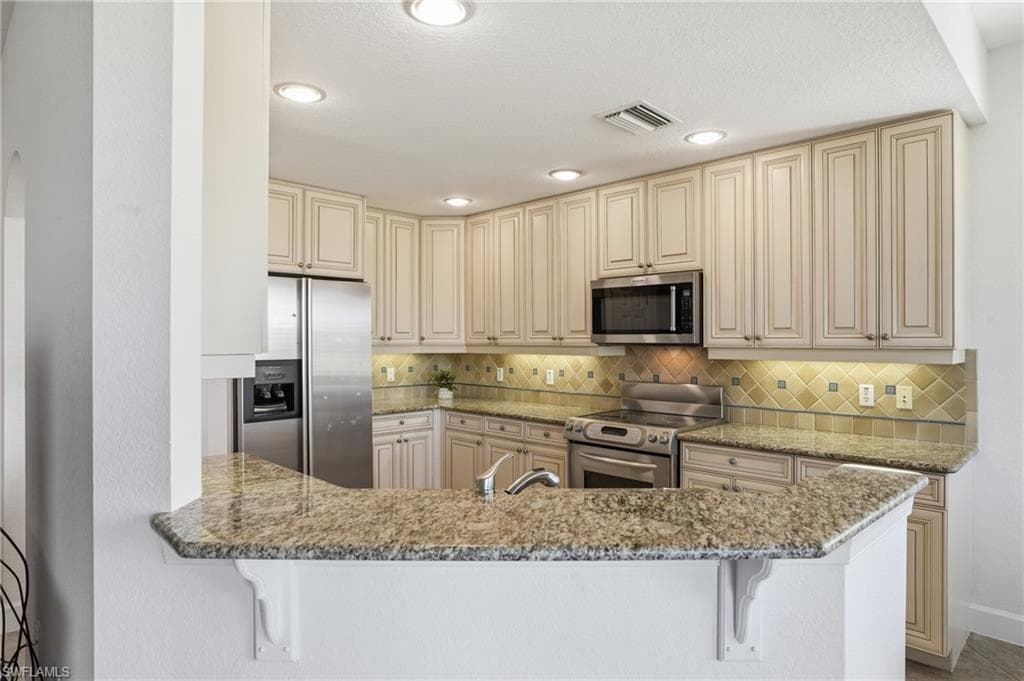 Kitchen featuring cream cabinets, stainless steel appliances, light stone countertops, a kitchen bar, and recessed lighting
