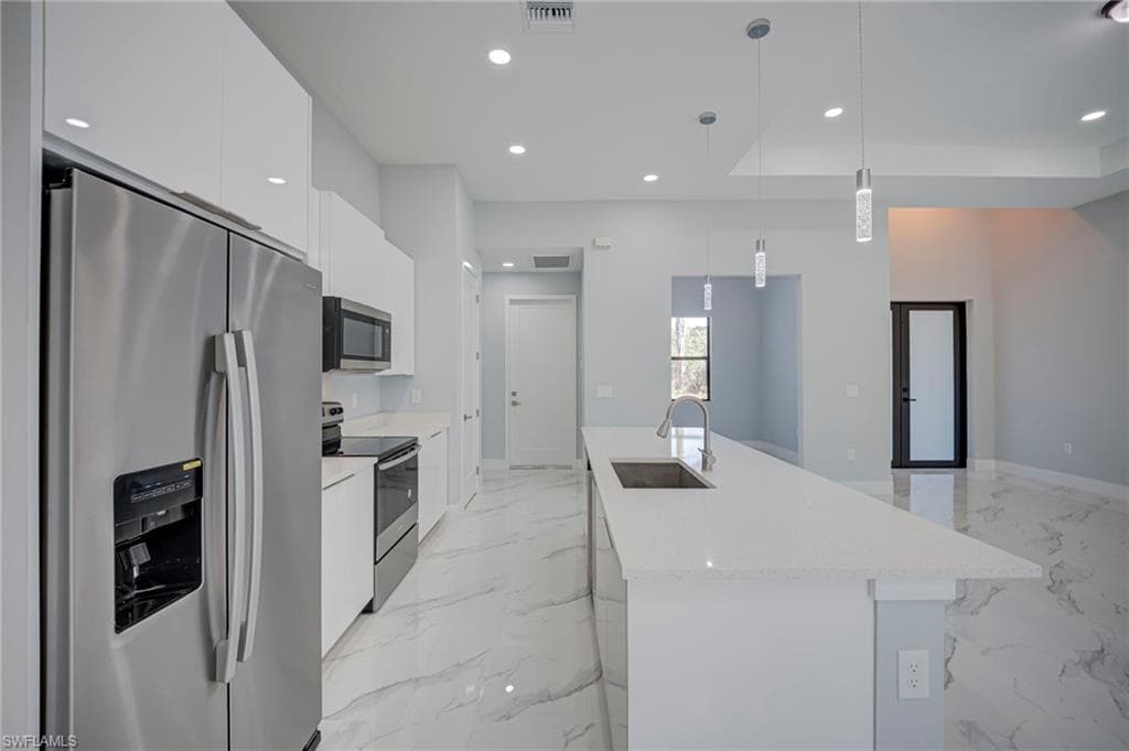 Kitchen featuring stainless steel appliances, an island with sink, white cabinets, decorative light fixtures, and light stone countertops