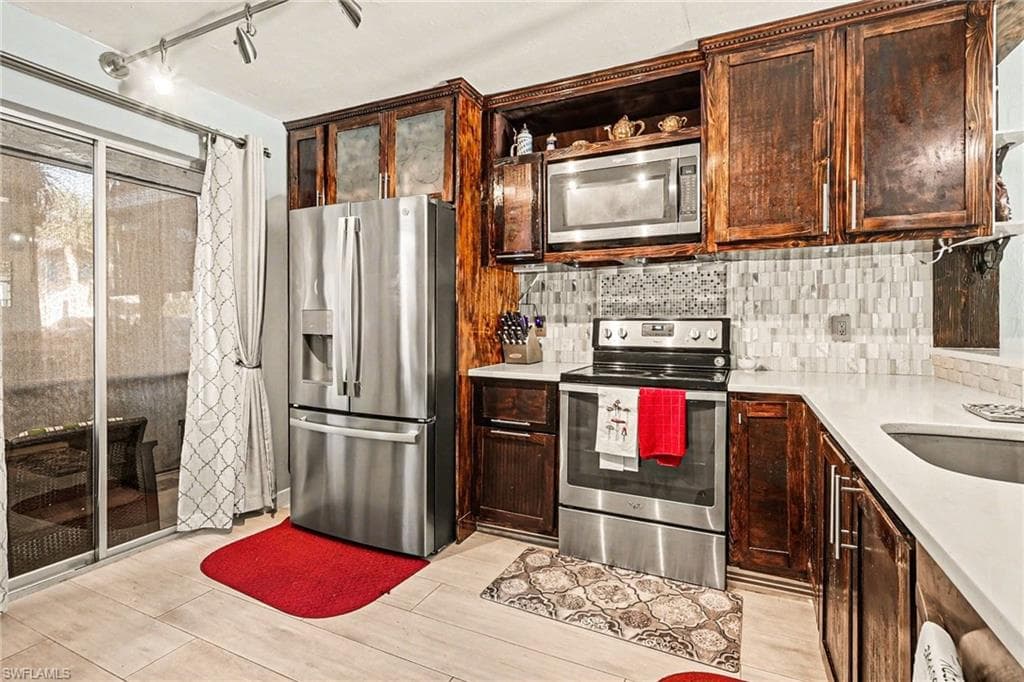 Kitchen with stainless steel appliances, open shelves, rail lighting, dark wood finish cabinetry, and light stone counters