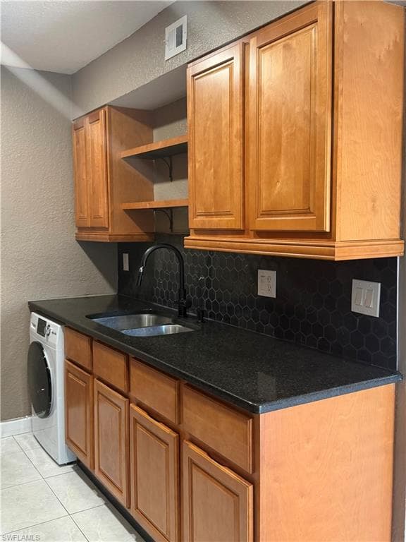 Functional utility space featuring wood-finish cabinetry, dark stone countertops, a stainless steel sink with a matte black faucet, and a black hexagonal tile backsplash