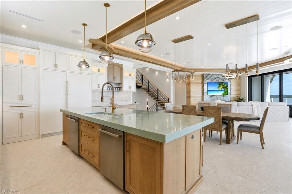 Open-concept kitchen and dining area featuring a substantial island with wood cabinetry and waterfall-edge countertop, stainless steel appliances, white perimeter cabinetry with brass hardware, exposed beam ceiling detail, pendant lighting, and expansive windows with natural light throughout the space - Virtually Edited Image