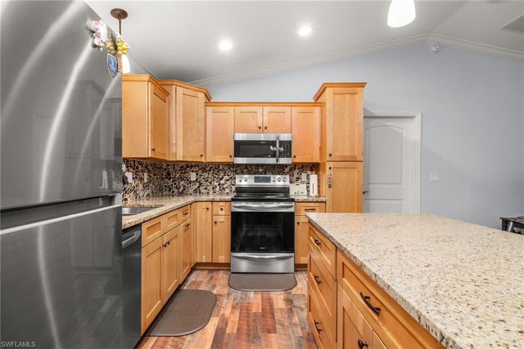 Kitchen with appliances with stainless steel finishes, tasteful backsplash, light stone countertops, vaulted ceiling, and light brown cabinetry