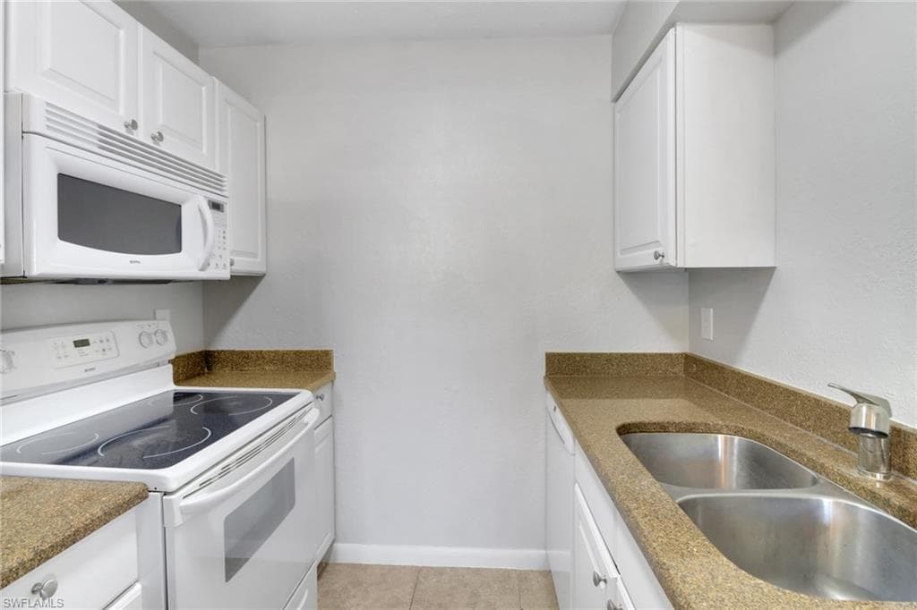 Kitchen featuring a sink, white cabinetry, white appliances, light tile patterned flooring, and baseboards