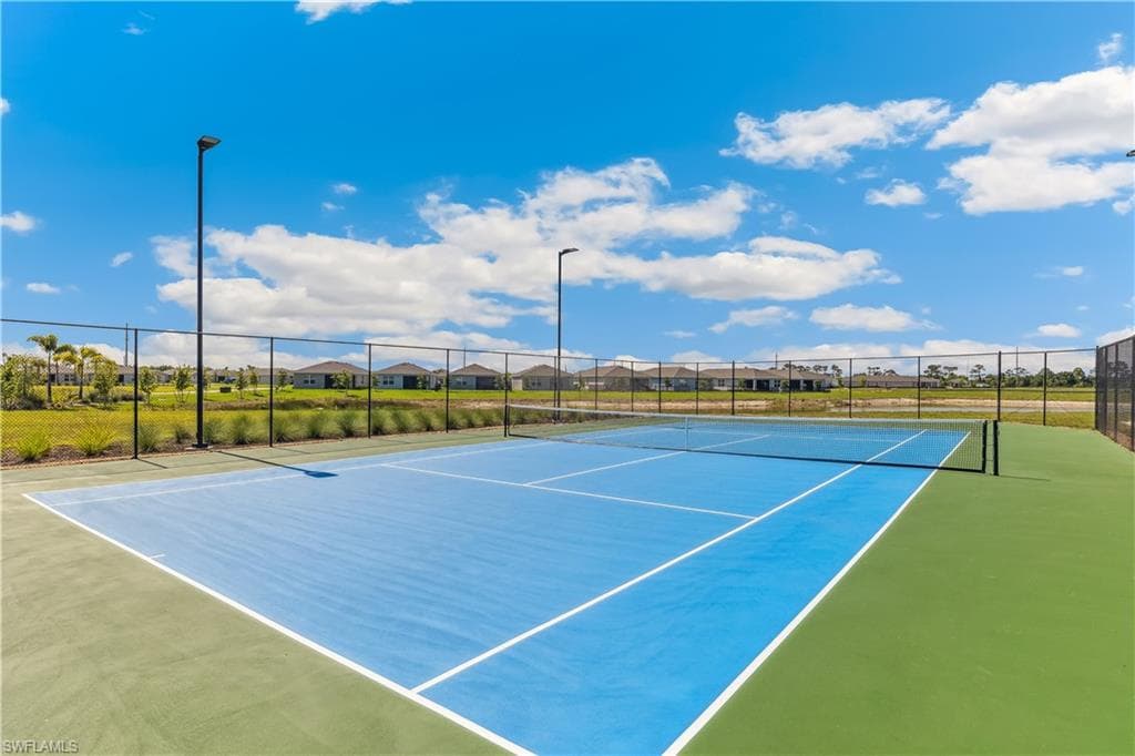 Outdoor tennis court featuring a blue playing surface and green surrounds, enclosed by a fence