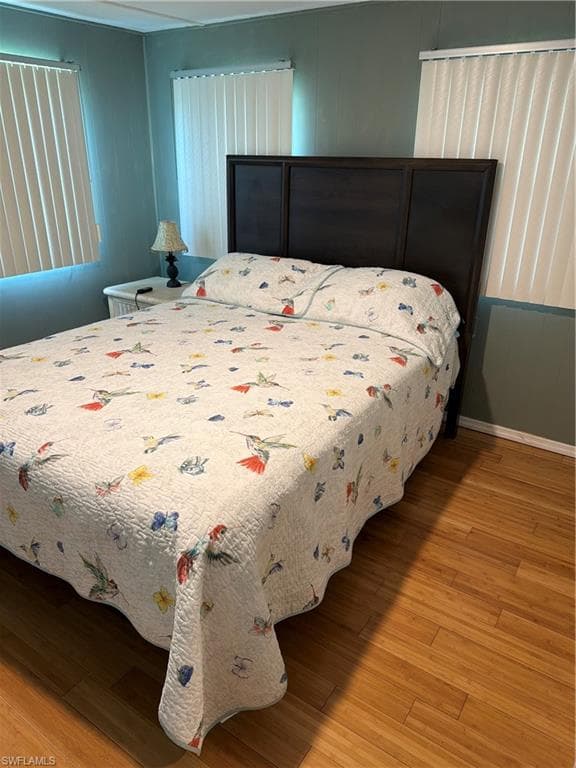 Bedroom featuring wood-finish flooring, a dark-toned headboard, and two windows with vertical blinds