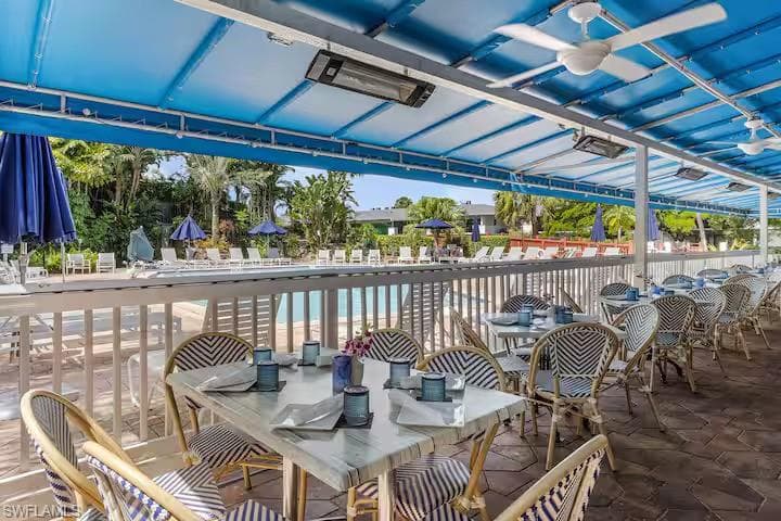 Outdoor dining area featuring a blue canopy with ceiling fans and heaters, overlooking a swimming pool area with lounge chairs and umbrellas, and surrounded by tropical landscaping