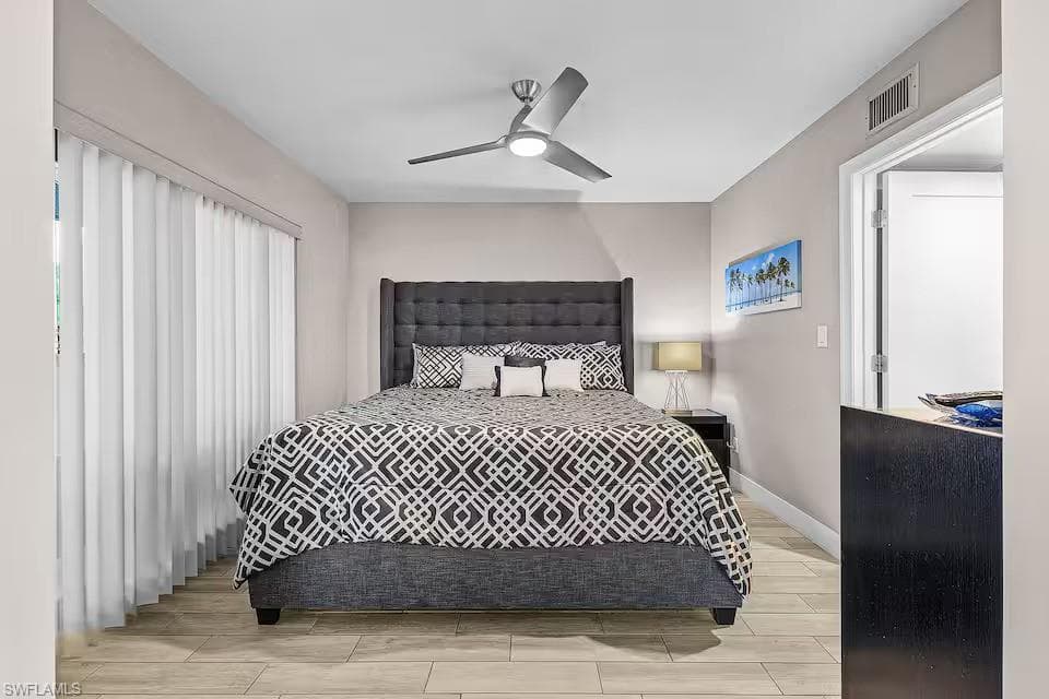 This sleeping area features light-toned flooring, a ceiling fan, and vertical blinds