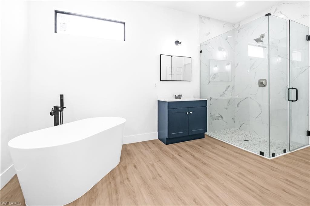 Contemporary bathroom featuring a freestanding soaking tub with matte black fixtures, a spacious glass-enclosed shower with white marble-patterned tile, a dark blue vanity with a white countertop and chrome faucet, wood-finish flooring, and a horizontal transom window