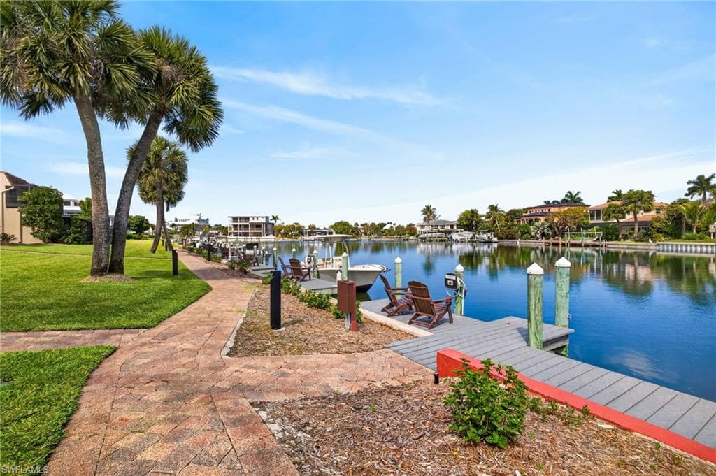 Waterfront property featuring a community dock with pilings, a paved pedestrian pathway, and mature palm trees