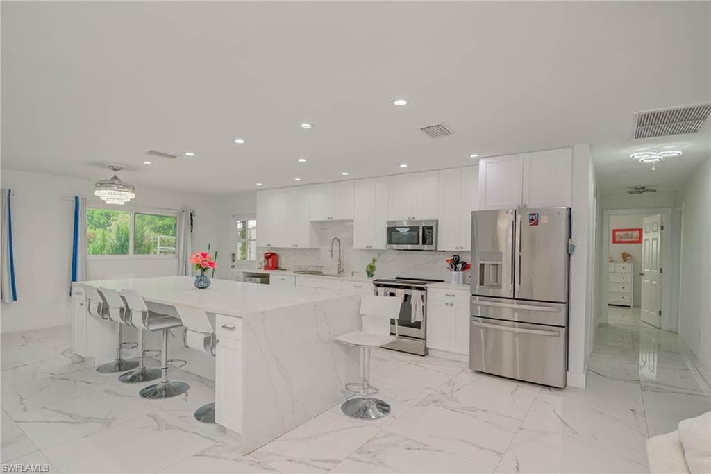 Spacious kitchen featuring an extended waterfall island, white cabinetry, stainless steel appliances, recessed lighting, and large format marble-look tile flooring