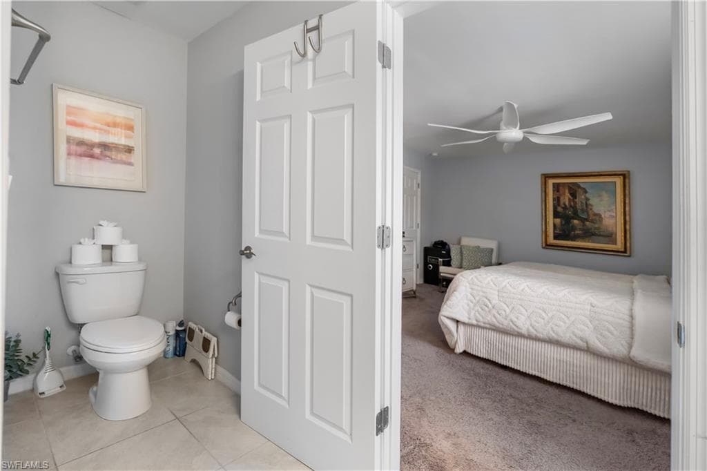 Bathroom featuring tiled flooring and a toilet, with a doorway opening to a carpeted room featuring a ceiling fan