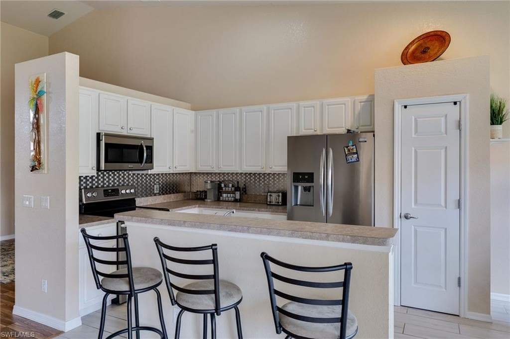 Kitchen featuring white cabinetry, stainless steel appliances, a black and white tiled backsplash, light-toned countertops, and a breakfast bar