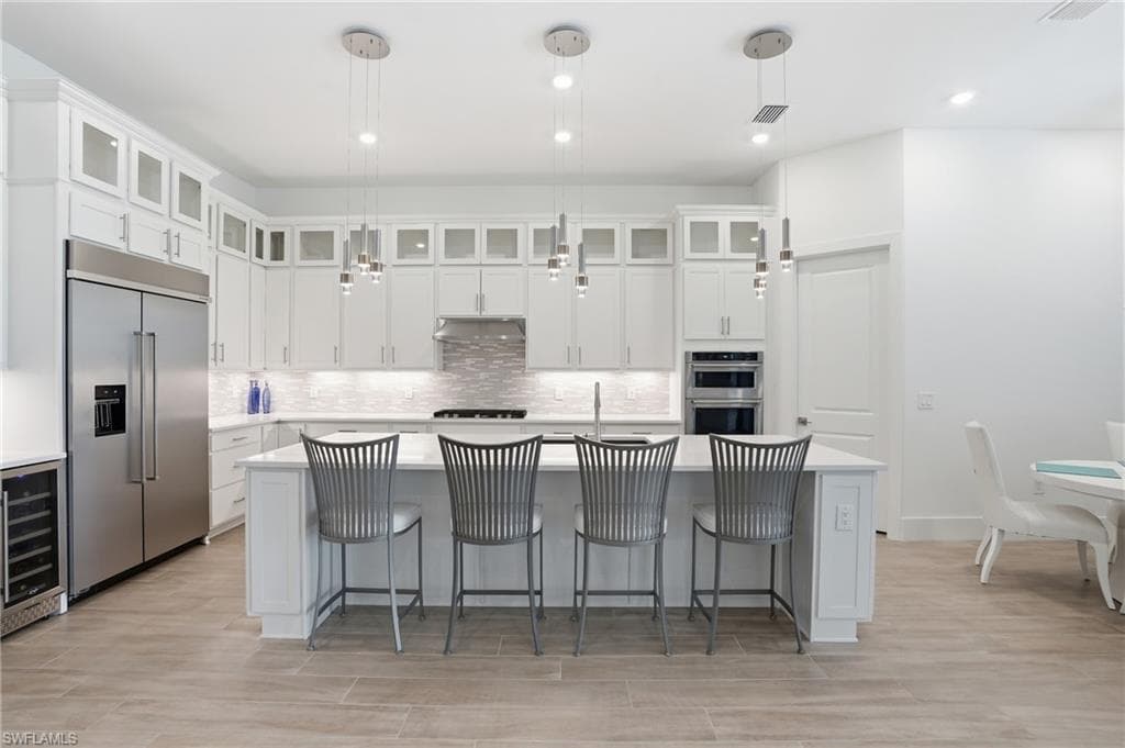 Gourmet kitchen featuring a central island with seating, stainless steel appliances, extensive white cabinetry with upper glass-front accents, a mosaic tile backsplash, and recessed and pendant lighting