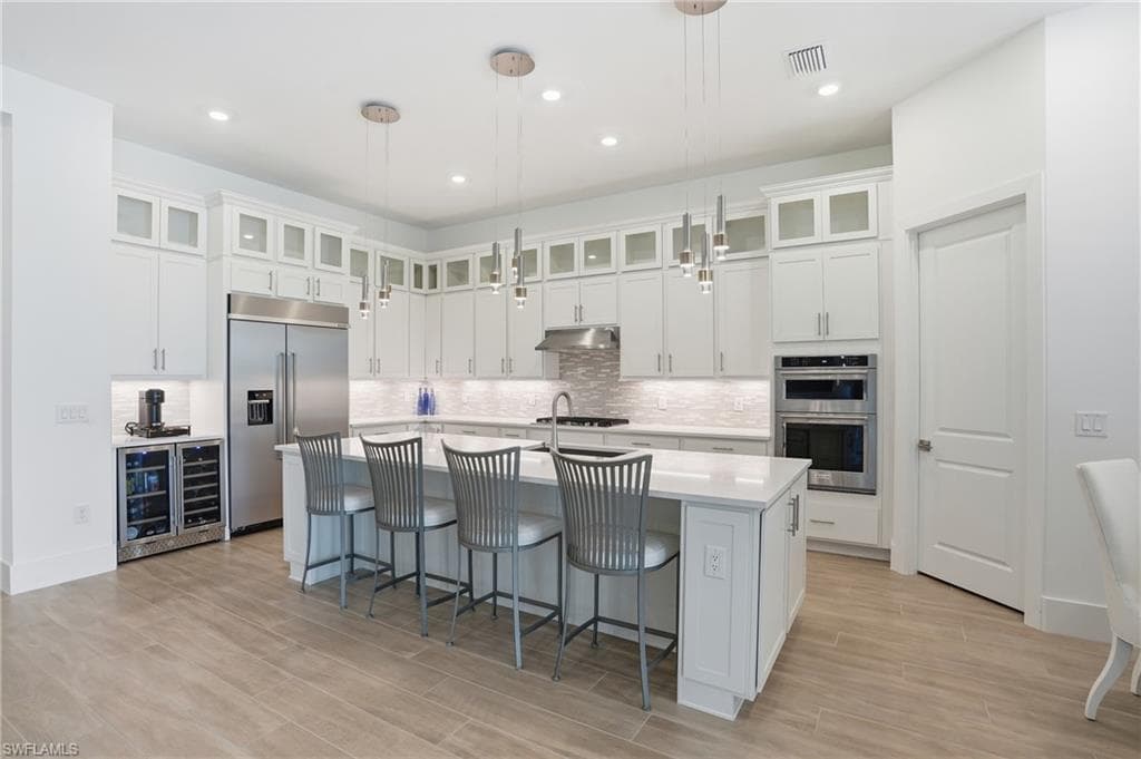 Spacious kitchen featuring an expansive island with seating, stainless steel appliances, white cabinetry with glass-front uppers, a tiled backsplash, and wood-finish flooring