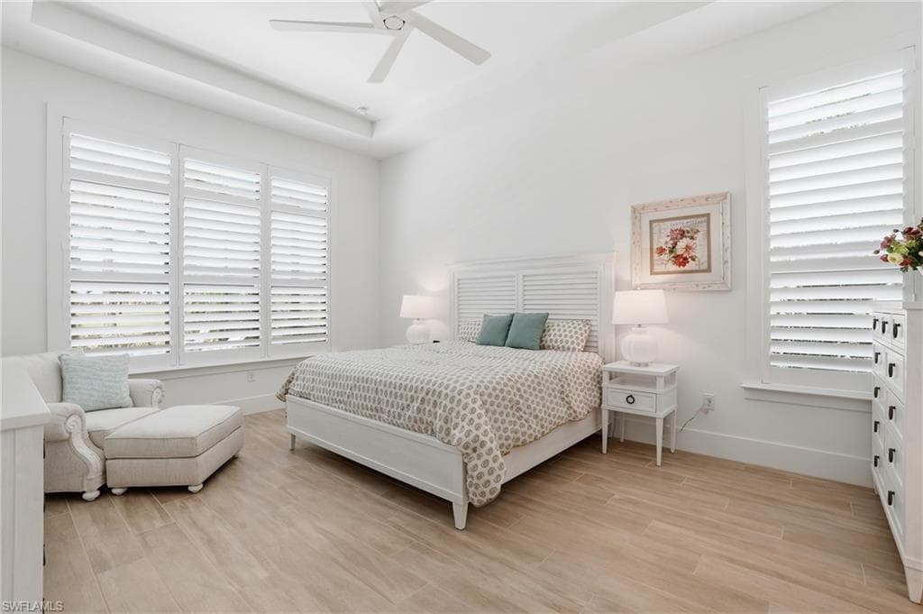 Spacious bedroom featuring wood-finish flooring, white plantation shutters, a tray ceiling, and a ceiling fan