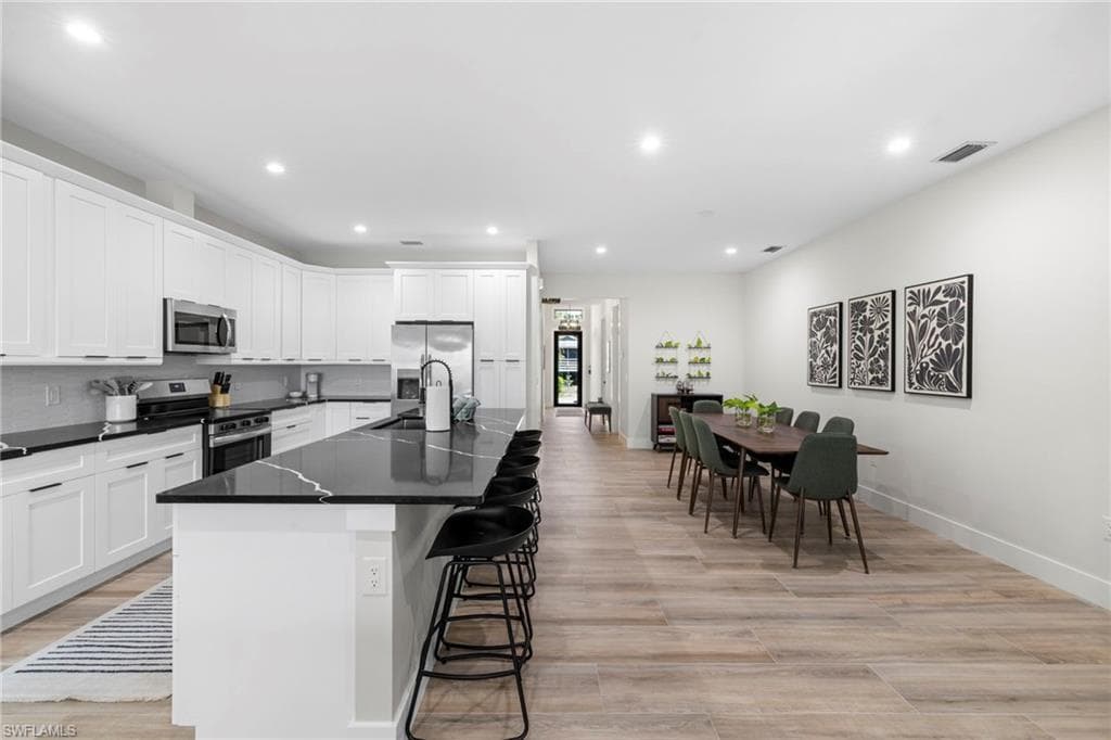 Spacious kitchen and dining area featuring an expansive island with seating, white shaker cabinetry, stainless steel appliances, dark countertops with light veining, and light wood-finish flooring