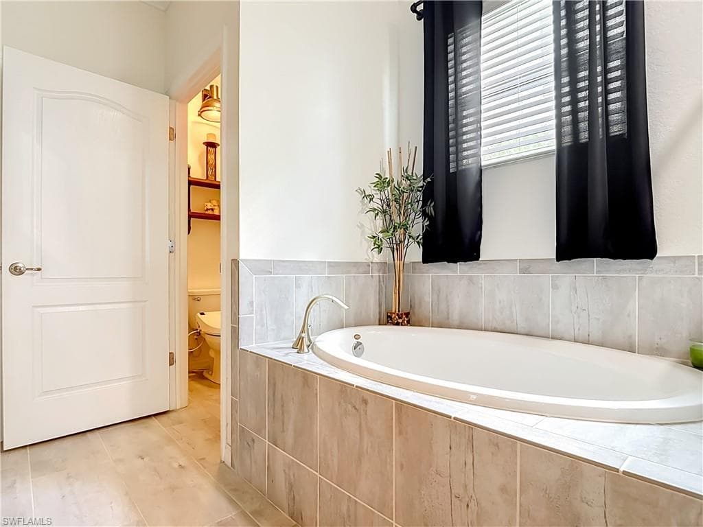 Built-in soaking tub featuring light-toned tile surround, a brushed nickel faucet, and a window with blinds