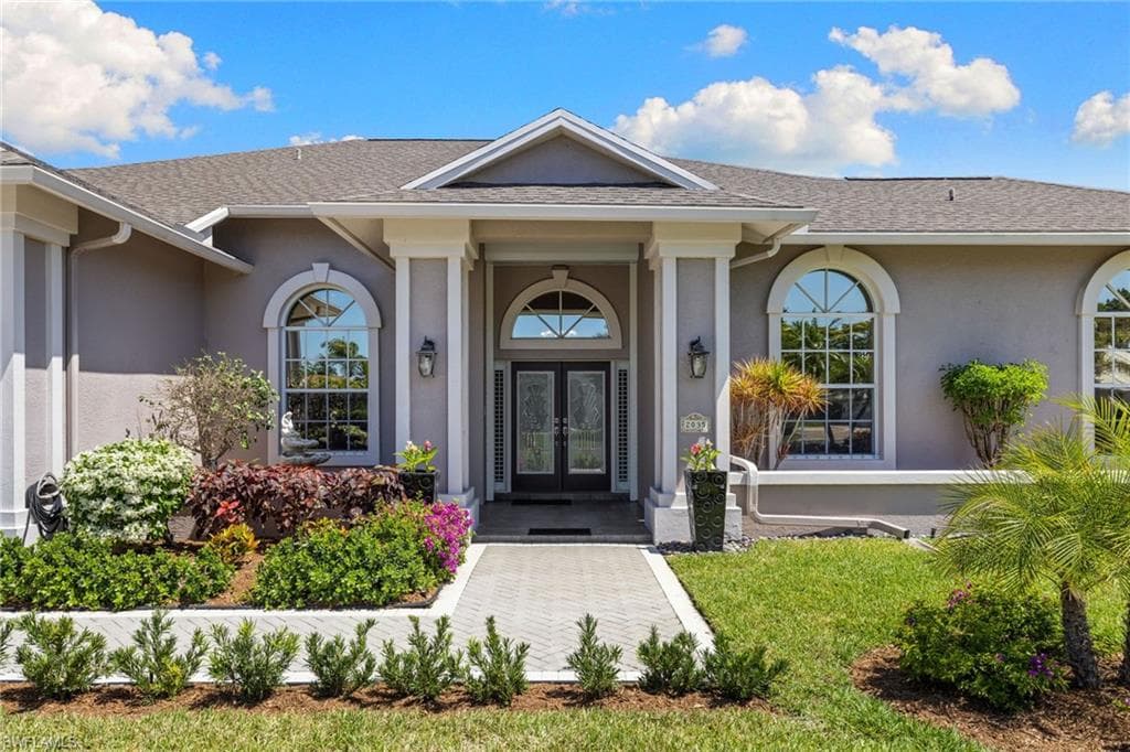 Exterior entry featuring a covered portico with substantial columns, arched windows, a double front door, a paver walkway, and mature landscaping