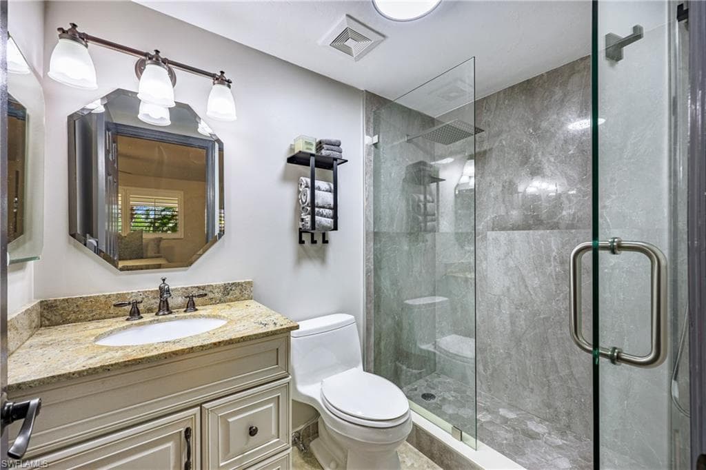 Bathroom featuring a glass-enclosed shower with gray stone-finish wall tile, a white vanity with a light-toned countertop, an octagonal mirror, a three-light vanity fixture, and a white toilet