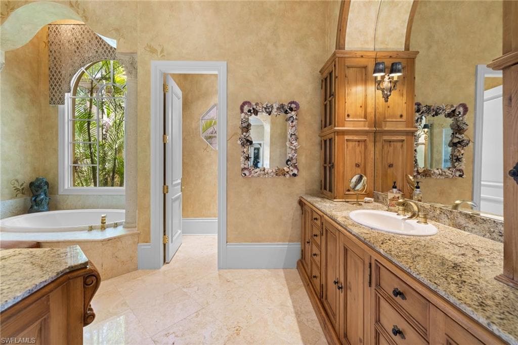 Bathroom featuring a built-in jetted tub under an arched window, a long vanity with dual sinks, wood cabinetry, and stone countertops