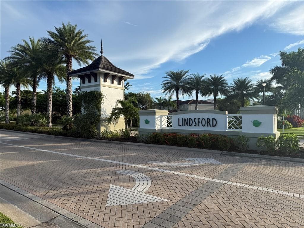 Paver brick entrance with a decorative tower featuring a dark roof and finial, alongside a community sign with a patterned accent and tropical landscaping, including multiple mature palm trees