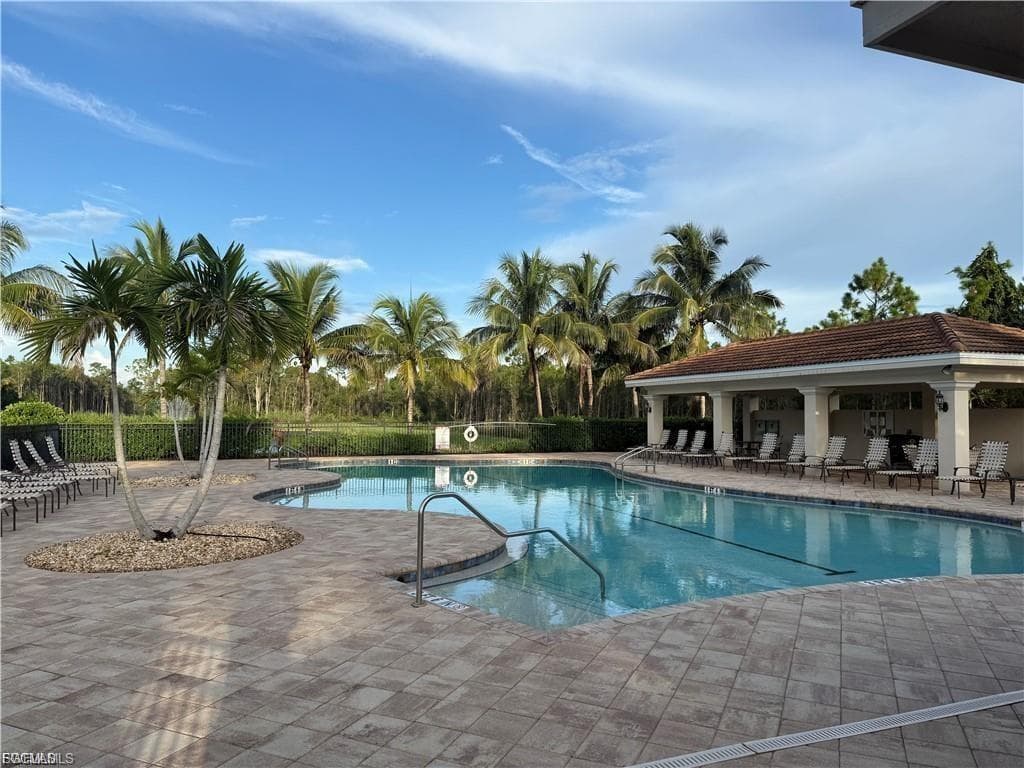 Resort-style pool area featuring a free-form swimming pool, paver deck, covered cabana with columns, lush palm trees, and a surrounding privacy hedge