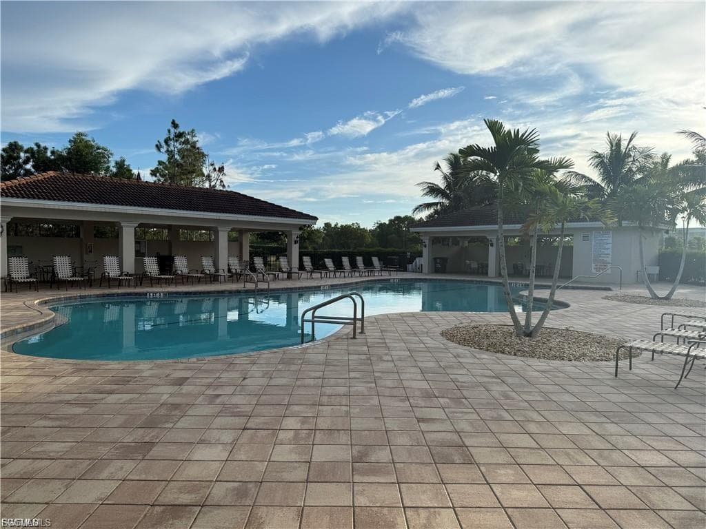 Resort-style pool area featuring a large swimming pool, expansive paver deck, covered lounge area with columns, and mature palm trees