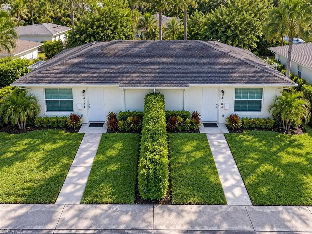Duplex property featuring a shingled roof, white exterior, front entryways, and manicured landscaping with mature shrubs and palm trees