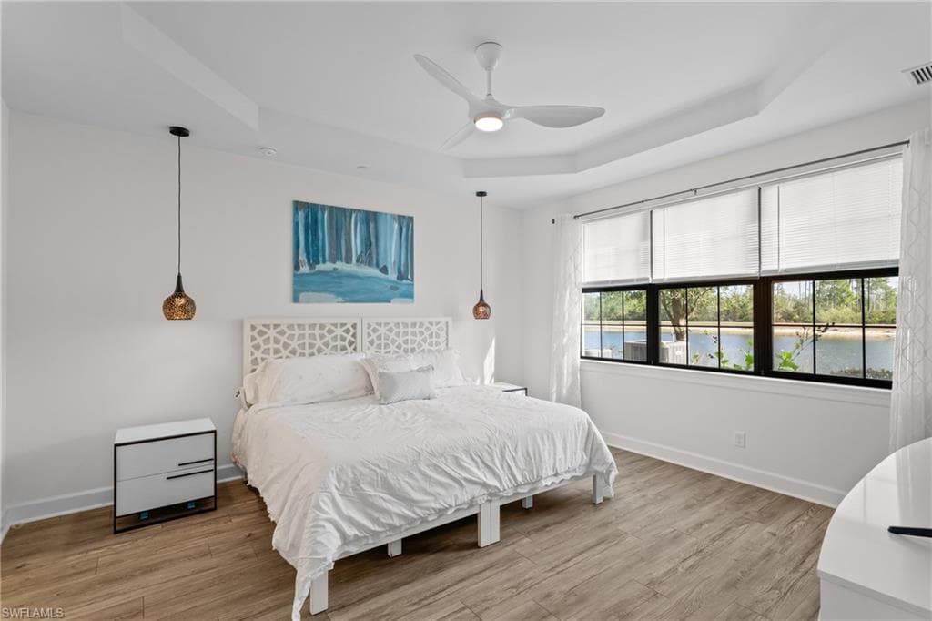 Bedroom featuring a raised ceiling, light wood-style flooring, ceiling fan, and a water view