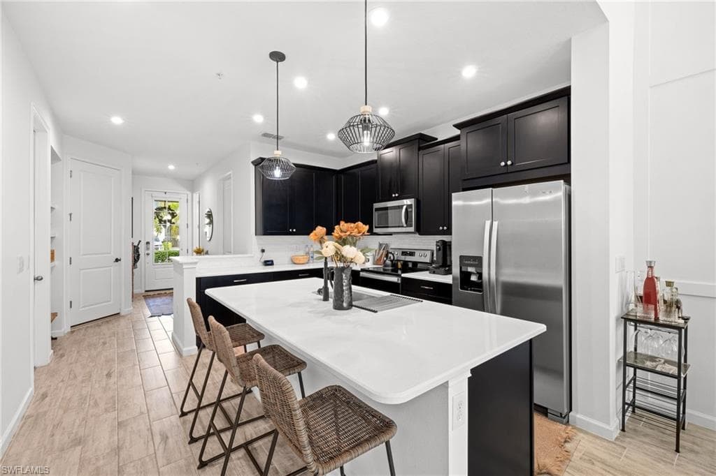 Modern kitchen featuring a spacious island with a white countertop, dark cabinetry, stainless steel appliances, and two pendant light fixtures