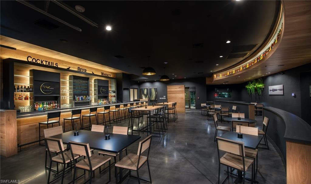 Spacious commercial interior featuring a full-service bar with built-in cabinetry and task lighting, polished concrete flooring, a curved wood-finish wall panel with integrated shelving, and various seating arrangements including high-top tables and standard dining tables