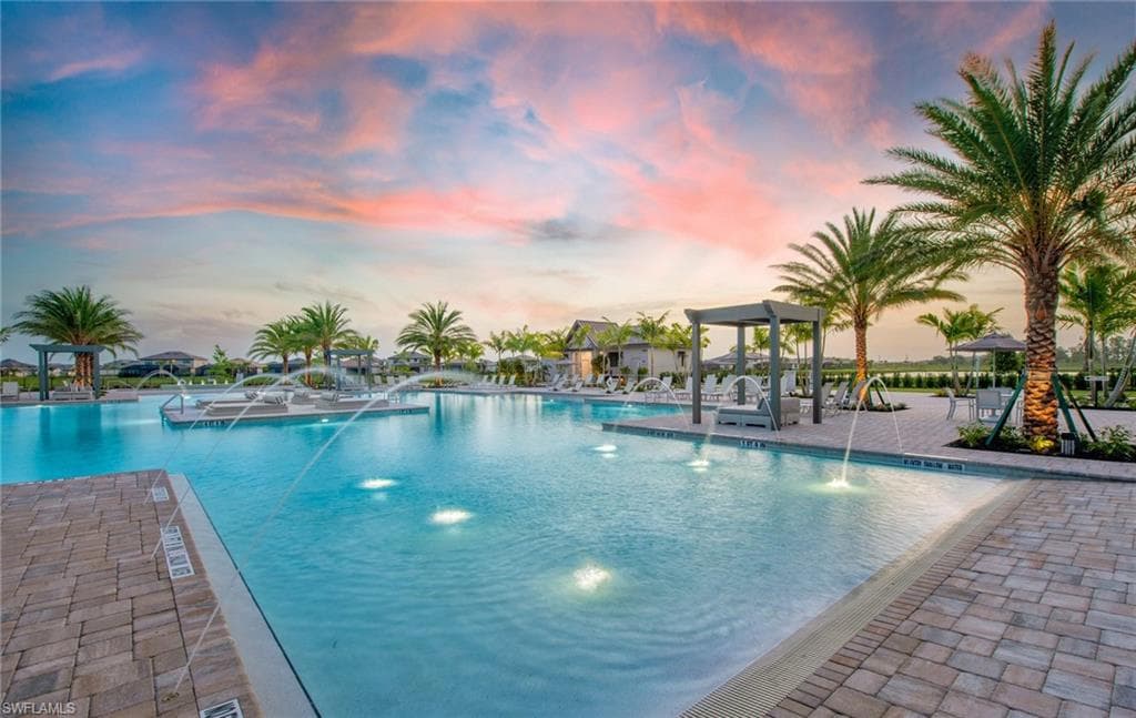 Resort-style pool area featuring a large swimming pool with fountains, surrounding paver decking, mature palm trees, multiple pergolas, and lounge seating