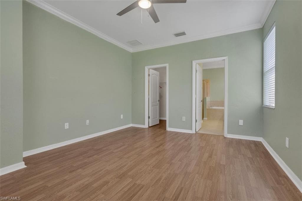 Unfurnished bedroom with a spacious closet, crown molding, light wood-style floors, ceiling fan, and ensuite bath