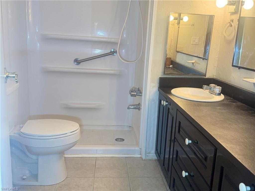 Bathroom featuring a shower stall with integrated shelving and a grab bar, a white toilet, a dark vanity with an integrated sink, a dark countertop, and tiled flooring